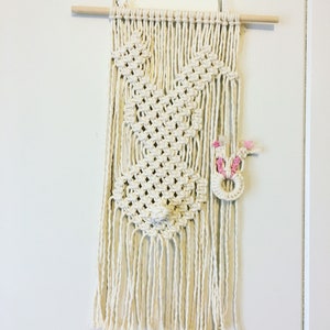 May include: A white macrame wall hanging with a bunny design and pink accents. The hanging is attached to a wooden dowel rod.