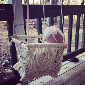May include: A baby in a white macrame swing with a pink bow in their hair. The swing is hanging from a wooden porch railing.