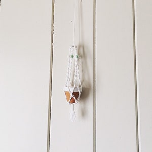 May include: White macrame plant hanger with a small terracotta pot hanging from it. The hanger is attached to a white wall.