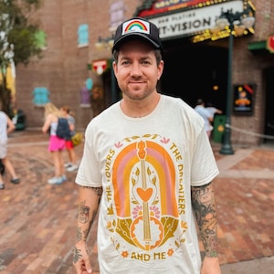 May include: A person wearing a white t-shirt with a colorful graphic of a guitar, rainbow, and text that reads "The Lovers, The Dreamers, and Me." The person is also wearing a black baseball cap with a rainbow design.