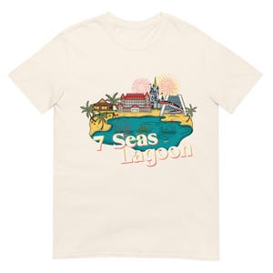 May include: Cream-colored t-shirt featuring a colorful graphic of a lagoon with buildings, palm trees, and fireworks. The text "7 Seas Lagoon" is printed in a retro font, evoking a vintage travel aesthetic.