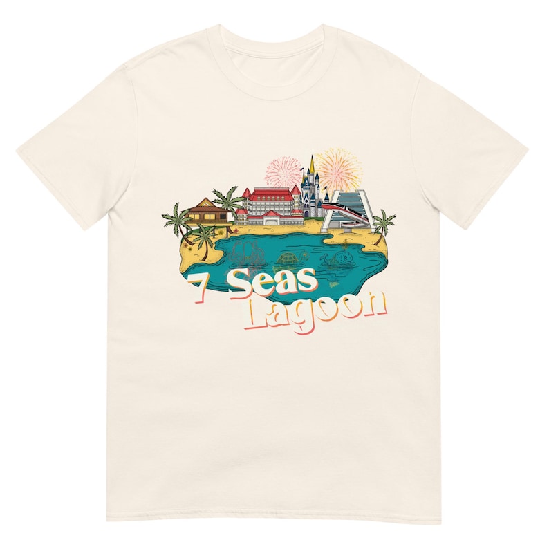May include: Cream-colored t-shirt featuring a colorful graphic of a lagoon with buildings, palm trees, and fireworks. The text "7 Seas Lagoon" is printed in a retro font, evoking a vintage travel aesthetic.