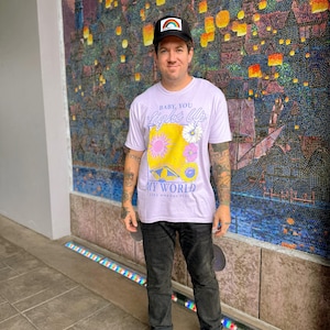 May include: A person wearing a black baseball cap with a rainbow design, a light purple t-shirt with the text "BABY, YOU LIGHT UP MY WORLD LIKE NOBODY ELSE", black jeans, and white trainers. The background features a colorful mosaic.