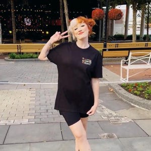 Puede incluir: Una persona con una camiseta negra con el texto "It's the Business, Baby!" y pantalones cortos negros. La persona lleva sandalias negras. El fondo incluye árboles, bancos y un edificio.