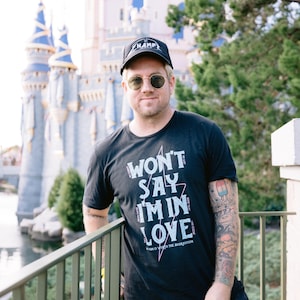 May include: A person wearing a black t-shirt with the text "WON'T SAY I'M IN LOVE" and a black baseball cap. The person is wearing sunglasses and standing in front of a castle and trees.