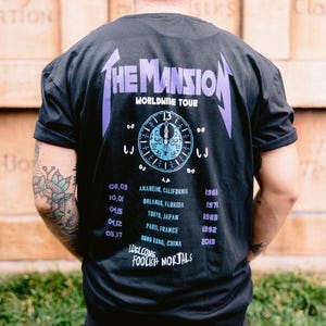 May include: A black t-shirt featuring a graphic on the back with the text THE MANSION WORLDWIDE TOUR, a clock face, a list of cities including Anaheim, Orlando, Tokyo, Paris, and Hong Kong, and the text WELCOME FOOLISH MORTALS in purple and blue.