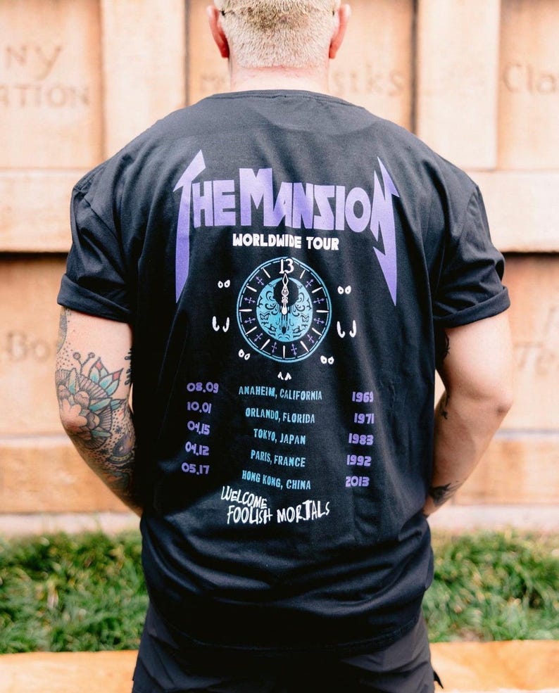May include: A black t-shirt featuring a graphic on the back with the text THE MANSION WORLDWIDE TOUR, a clock face, a list of cities including Anaheim, Orlando, Tokyo, Paris, and Hong Kong, and the text WELCOME FOOLISH MORTALS in purple and blue.