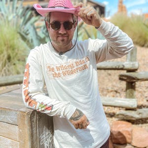 May include: A person wearing a pink sequined cowboy hat and sunglasses. They are wearing a long-sleeved, off-white shirt with the text "The Wildest Ride in the Wilderness" and a colourful design on the sleeve.