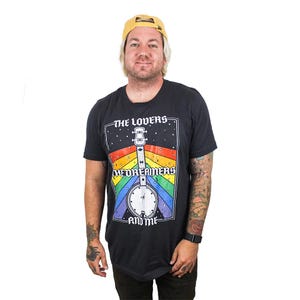 May include: Dark gray t-shirt with a graphic of a banjo and the text "THE LOVERS, MEDREAMERS, AND ME" in a rectangular design with a rainbow background. The banjo is white with a clock face. The shirt is short-sleeved.