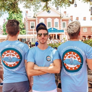 Puede incluir: Tres personas con camisetas azul claro con un diseño gráfico circular con un águila, estrellas y el texto "A Salute to All Nations, But Mostly America". Las camisetas tienen un pequeño logotipo en la parte delantera. El fondo incluye un edificio con la palabra "PRESIDENTS".