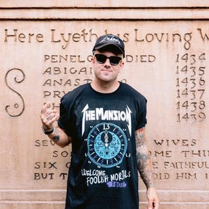 May include: A black short-sleeve t-shirt featuring a white graphic of a clock face with the text The Mansion, 13, and Welcome Foolish Mortals Tour. The shirt is paired with a black baseball cap and black sunglasses, creating a casual ensemble.