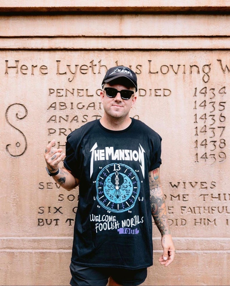 May include: A black short-sleeve t-shirt featuring a white graphic of a clock face with the text The Mansion, 13, and Welcome Foolish Mortals Tour. The shirt is paired with a black baseball cap and black sunglasses, creating a casual ensemble.