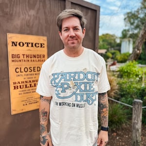 May include: A cream-colored t-shirt with the text "Pardon Pixie Dust" in blue and the phrase "I'm working on myself." The shirt is worn by a person standing in front of a wooden structure and a sign that reads "Big Thunder Mountain Railroad Closed."