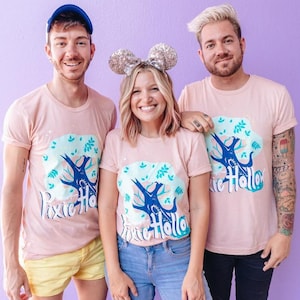 Puede incluir: Tres personas con camisetas rosas claras a juego con un diseño de árbol azul y verde y las palabras "Pixie Hollow". Una persona lleva una gorra azul, otra orejas de ratón con lentejuelas. El fondo es de color púrpura liso.