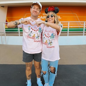 May include: Two people wearing matching light pink t-shirts with the text "There's A Great Big Beautiful Tomorrow" and a whimsical illustration. The person on the right is wearing a Minnie Mouse ear headband. The background is an orange and white wall.