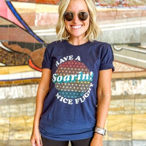 May include: Navy blue t-shirt with the text "Have a Soarin'! Nice Flight" in a colorful, circular design. The shirt is worn by a person wearing sunglasses and a watch.