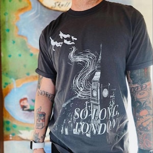 May include: Dark grey t-shirt with a white graphic of Big Ben and the words "SO LONG, LONDON." The design includes stylized clouds and birds. The shirt has short sleeves and a crew neck.