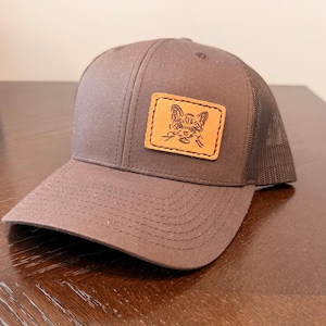 May include: A brown trucker cap with a mesh back and a curved peak. The cap has a rectangular leather patch with a cat face design. The cap is made of a durable fabric and is suitable for everyday wear.