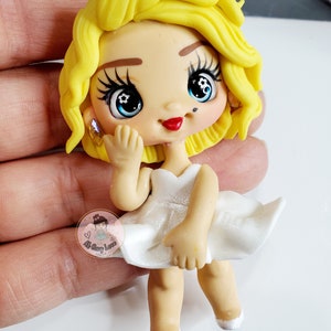 May include: A handmade figurine of a woman with blonde hair, red lips, and blue eyes, wearing a white dress. The figurine has a small beauty mark and is holding up her hand. A small logo is visible.