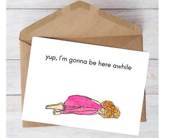 May include: A white greeting card with the text "yup, I'm gonna be here awhile" and an illustration of a person lying down. The card is inside a brown envelope, set against a white wood-grain background.