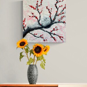 Op de afbeelding: Een schilderij van een gestileerde kersenboom met rode bloesems op een witte achtergrond. Het schilderij hangt aan een muur boven een vaas met zonnebloemen.