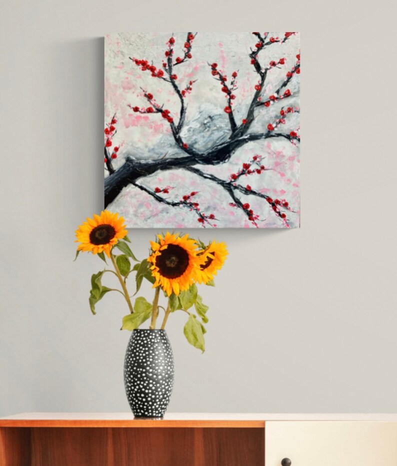 Op de afbeelding: Een schilderij van een gestileerde kersenboom met rode bloesems op een witte achtergrond. Het schilderij hangt aan een muur boven een vaas met zonnebloemen.