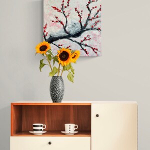 Op de afbeelding: Een witte kast met een bovenste houten plank en een enkele lade. De kast heeft een gesloten deur aan de rechterkant. Een vaas met zonnebloemen staat op de bovenste plank, en twee mokken met zwarte en witte stippen staan op de plank. Een schilderij van een bloeiende boom met rode bloemen hangt aan de muur achter de kast.