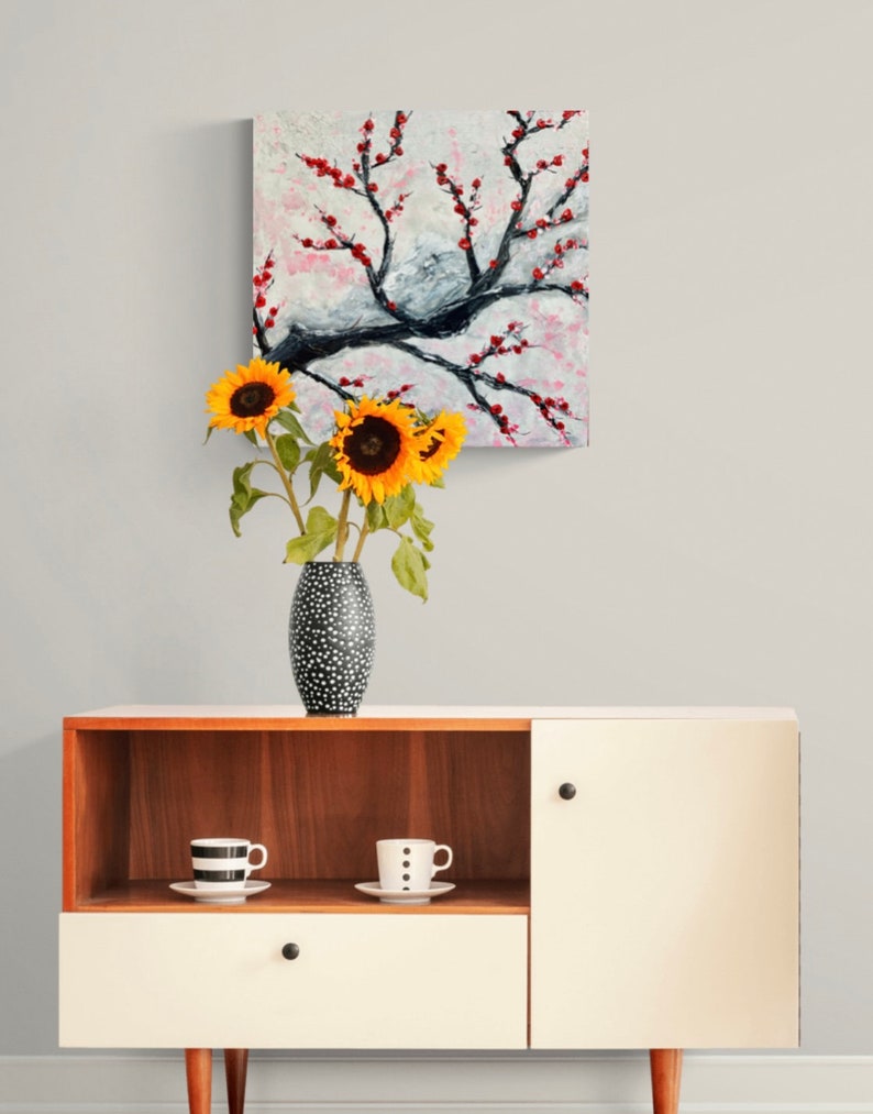 Op de afbeelding: Een witte kast met een bovenste houten plank en een enkele lade. De kast heeft een gesloten deur aan de rechterkant. Een vaas met zonnebloemen staat op de bovenste plank, en twee mokken met zwarte en witte stippen staan op de plank. Een schilderij van een bloeiende boom met rode bloemen hangt aan de muur achter de kast.