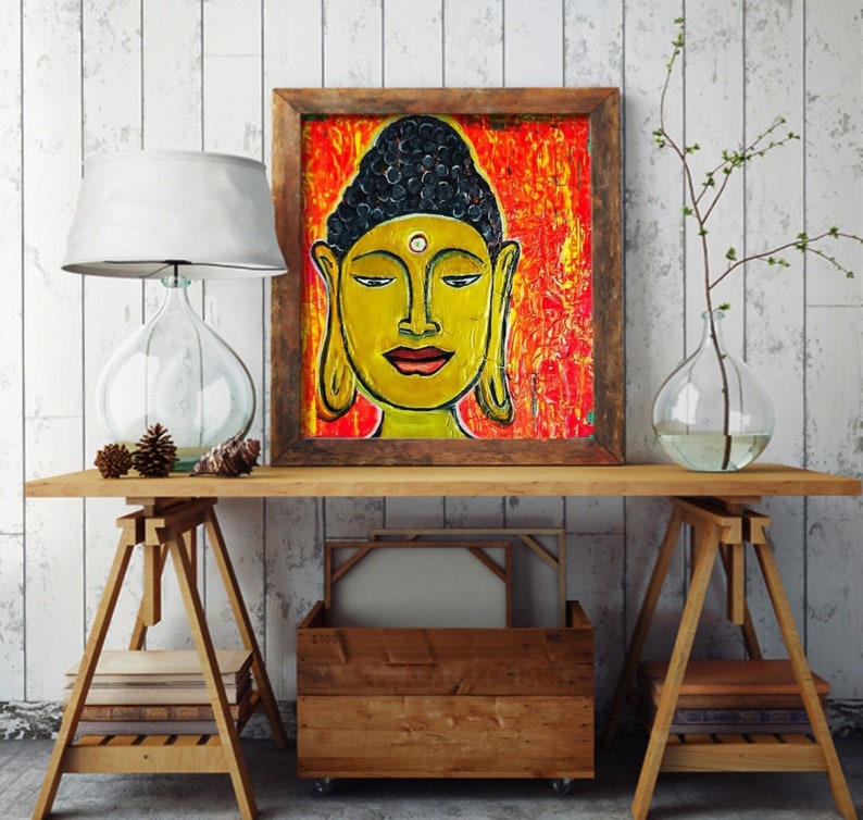 May include: A framed painting of a yellow Buddha with a black bun and a red dot on his forehead. The background is a vibrant orange and red abstract design.