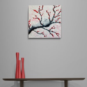 Op de afbeelding: Een schilderij van een tak met rode bloesems tegen een witte achtergrond. Het schilderij is ingelijst en hangt aan een grijze muur boven een tafel met drie rode vazen.