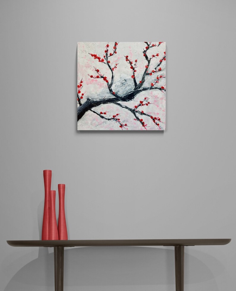 Op de afbeelding: Een schilderij van een tak met rode bloesems tegen een witte achtergrond. Het schilderij is ingelijst en hangt aan een grijze muur boven een tafel met drie rode vazen.