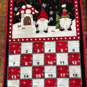 May include: A festive advent calendar with a red and white chequered pattern. The top panel shows gnomes, a mushroom house, and the text "from our gnome to yours". Numbers 1-24 are displayed across the calendar.
