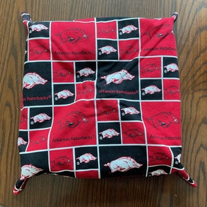 May include: Square fabric item with a repeating pattern of red and black squares. Each square features a white Razorback logo and the text "Arkansas Razorbacks". The item has four corner tabs.