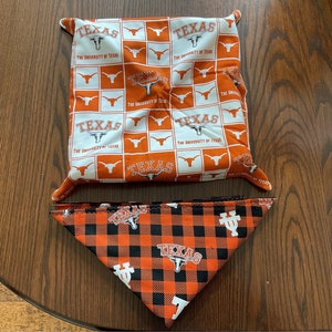 May include: A square fabric bowl cosy featuring a white and orange checkerboard pattern with the University of Texas logo and text, paired with a triangular pet bandana in an orange and black buffalo plaid print with Texas logos.