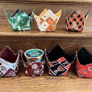 May include: Six fabric bowl cozies in various colors and patterns. The cozies are shaped like crowns and feature different designs, including a green and white pattern with the words "North Texas", a black and white plaid pattern, a red and white pattern with the words "Texas A&M", a black and white pattern with the words "Texas Tech", and a red and white pattern with the words "Texas A&M".