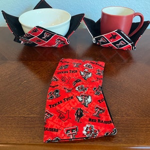 Peut inclure: Deux dessous de plat et de tasse en tissu rouge et noir avec un motif Texas Tech Red Raiders. Une serviette en tissu rouge et noir avec le même motif est pliée sur une table en bois.