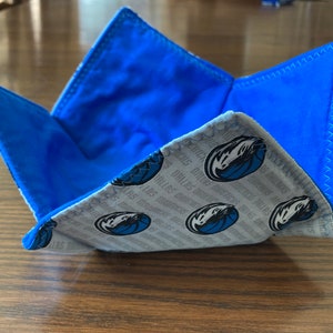 Peut inclure: Un bol en tissu bleu et gris avec un motif de logo des Dallas Mavericks. Le bol est plié et a une doublure bleue.
