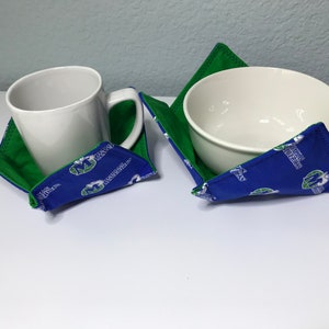 May include: Two fabric bowl and mug cozies with a green and blue design. The cozies feature a white logo with the text "Marquette Golden Eagles".