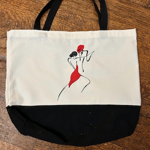 May include: A canvas tote bag with black handles and a black bottom. The main body is white, featuring a red and black line drawing of a couple dancing. The dancers are in a tango pose, with the woman in a red dress.