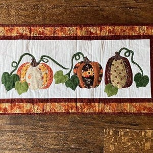 May include: A quilted table runner with a white background and a border of orange and brown fabric. Three pumpkins with green leaves are appliqued on the runner. The pumpkins are decorated with different patterns and colours.
