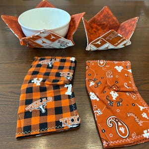 May include: Two fabric bowl cozies featuring orange and white Texas Longhorns patterns, one holding a white ceramic bowl. Below are two folded fabric handle covers, one with an orange and black buffalo plaid pattern and one with an orange paisley print.