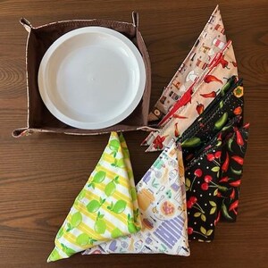 Puede incluir: Un juego de seis servilletas de tela con varios estampados de temática alimentaria, incluyendo limones, pimientos y cerezas. Las servilletas están dobladas en forma de triángulo y dispuestas alrededor de una cesta de tela marrón con un plato blanco en su interior.