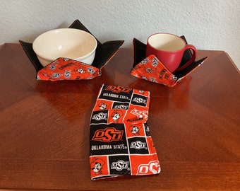 May include: Set of three items: a bowl cozy with a white bowl, a mug cozy with a red mug, and a folded item. All items feature a black and orange pattern with the text "Oklahoma State" and a cowboy logo.
