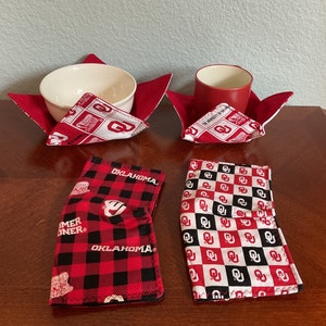 May include: Four red and white fabric pot holders with the Oklahoma Sooners logo. Two are folded into a square shape and two are flat. One of the flat pot holders has a black and red plaid pattern with the words "Sooner" and "Oklahoma".