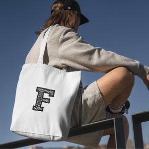 May include: A white tote bag with a black and white sketched letter "F" on the front. The bag is being carried by a person wearing a grey sweatshirt and shorts.