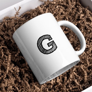 May include: White ceramic mug with a black hand-drawn letter "G" on the front. The mug is sitting in a box filled with brown packing paper.