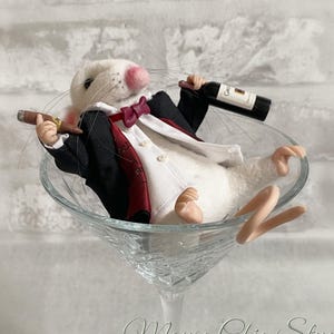 May include: A white felt mouse figurine wearing a black tuxedo jacket with a red vest and a maroon bow tie. The mouse holds a miniature cigar in one hand and a small wine bottle in the other, while reclining inside a clear glass martini glass.