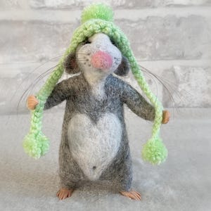 May include: A felted mouse figurine with a gray body, white chest in a heart shape, and a pink nose. It wears a green knit hat with long ear flaps and pom-poms. The figurine is standing on a light gray surface.
