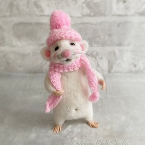 May include: A handmade white mouse figurine wearing a pink knitted hat and scarf. The mouse has long whiskers and small, detailed feet. The figurine is standing and smiling, with the text "Mouse Chic Studio" in the bottom right corner.
