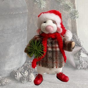 May include: A felt mouse figurine wearing a red and white Santa hat, a red scarf, and a brown plaid button-up jacket. The mouse holds a small green pine sprig and wears bright red shoes, standing upright with a pink nose and white fur.